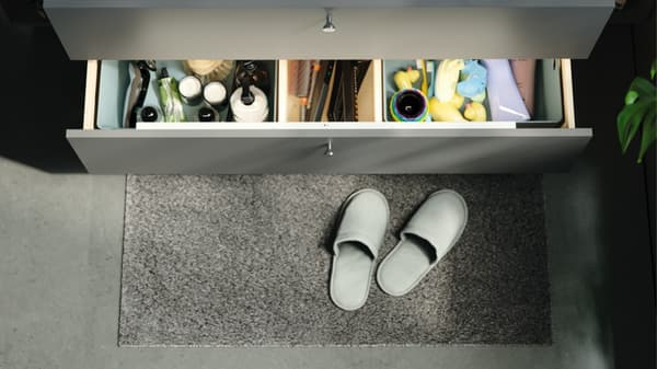 Des pantoufles blanches sur un tapis de bain gris, en partie sous un tiroir ouvert d'un meuble de salle de bain gris.