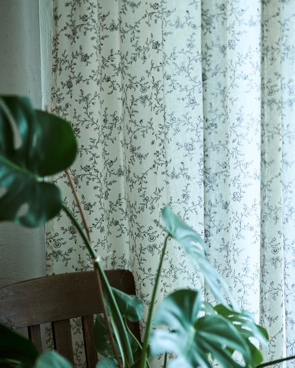 Des rideaux blancs avec un délicat imprimé floral noir accrochés à une fenêtre à côté d’une chaise de salle à manger et d’une plante MONSTERA.