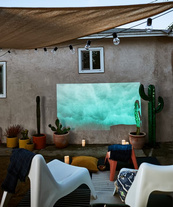 Des sièges blancs devant une image projetée sur un mur extérieur, sous une toile et une guirlande lumineuse, plus des bougies et des plantes en pot.