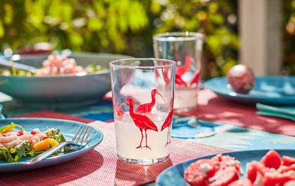 Des tables dressées dehors au soleil, avec de la vaisselle et des verres aux motifs et coloris estivaux. Des plats légers sont servis sur les assiettes.
