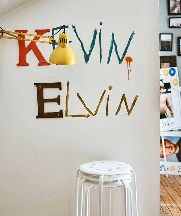 Des tabourets empilés à côté d’un mur sur lequel sont peints les noms Kevin et Elvin et une lampe de table jaune.