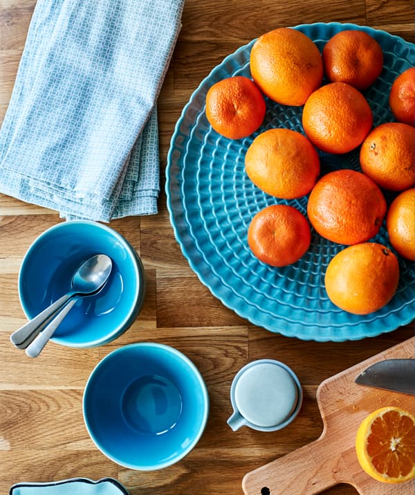 Des tasses, des serviettes et un plateau turquoise avec des mandarines, sur un plan de travail en bois à côté d’une planche à découper avec une moitié de mandarine.