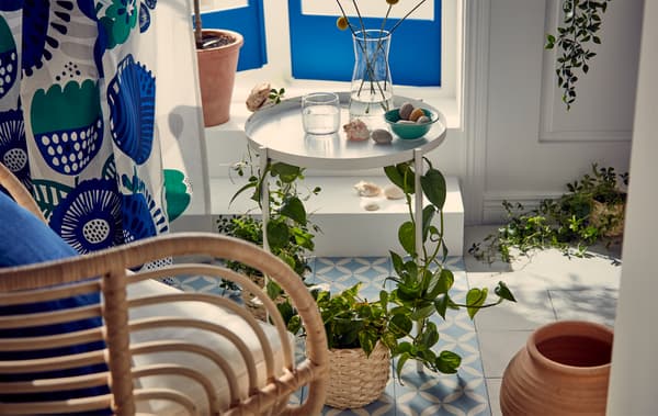 Des tons bleus et blancs dans un coin ensoleillé juste à l’intérieur d’une fenêtre donnant sur un balcon à la française. Un fauteuil en rotin à côté d’une table-plateau dont les pieds sont drapés de plantes.
