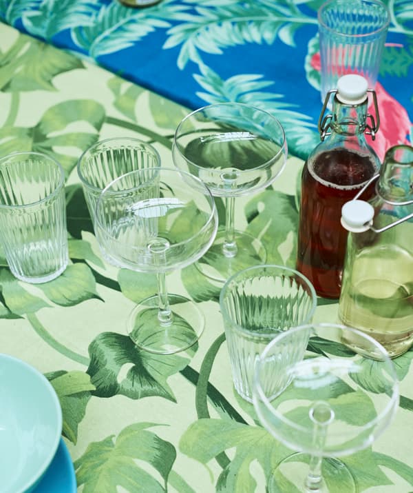 Des verres à champagne et à eau et des bouteilles en verre remplies sont posés sur une table recouverte d’un tissu à imprimé de feuilles vertes.
