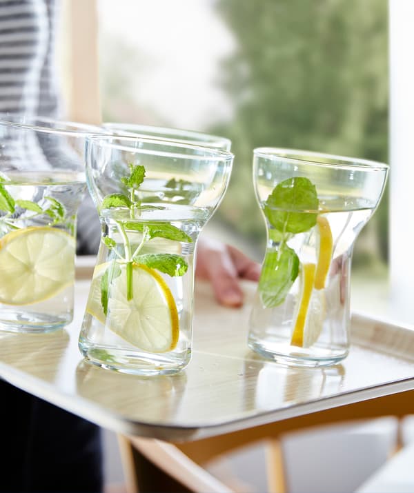 Des verres OMTÄNKSAM offrant une bonne prise, remplis d’eau, de tranches de citron et de brins de menthe, déposés sur un plateau antidérapant.