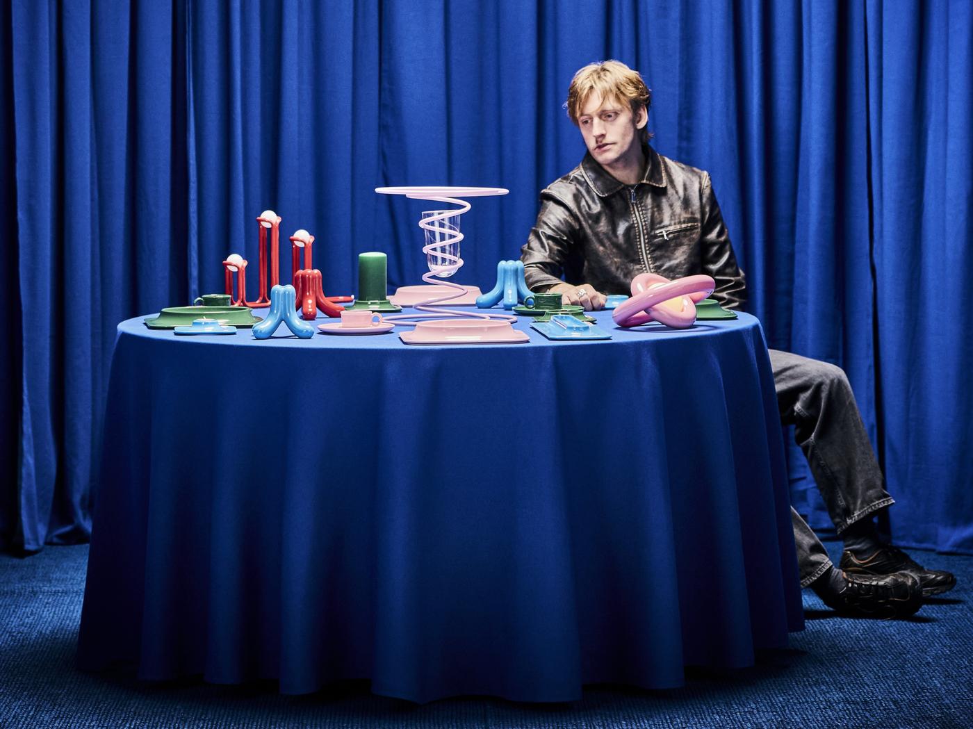 Designer Gustaf Westman an einem runden Tisch mit blauem Tischtuch, darauf seine 12- Designstücke