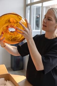 Designer Sabine Marcelis inspects a prototype of a VARMBLIXT Serving bowl with lid, glass/orange, with her assistant in her studio.