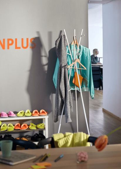 Desk with mug and notepad on it in the foreground with a shoe rack and coat hanger against a wall in the background