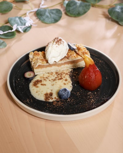 Dessert plate with a slice of apple pie topped with vanilla ice cream, poached pear, custard sauce, and blueberries, elegantly served on a black plate.
