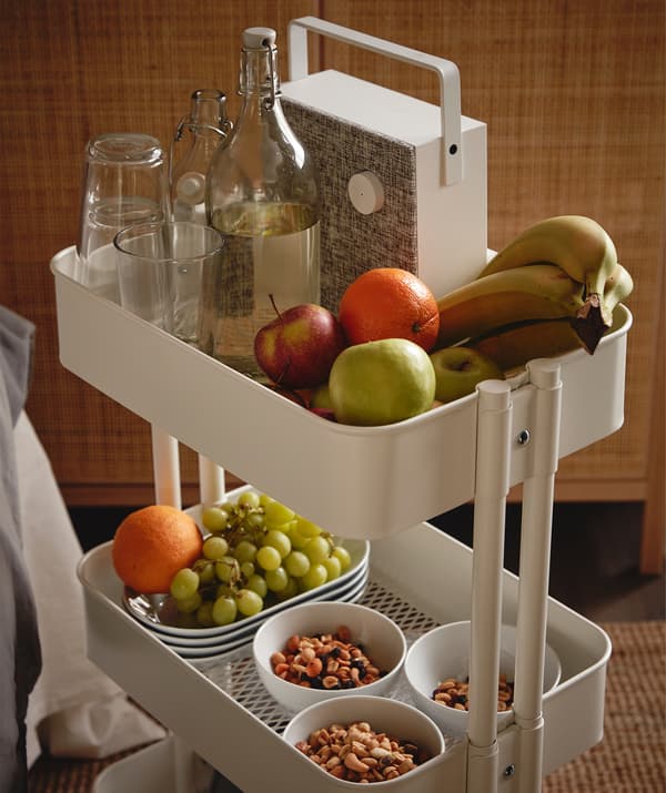 Desserte RÅSKOG remplie de différents en-cas et de fruits, une bouteille et des verres KORKEN et une enceinte Bluetooth ENEBY.