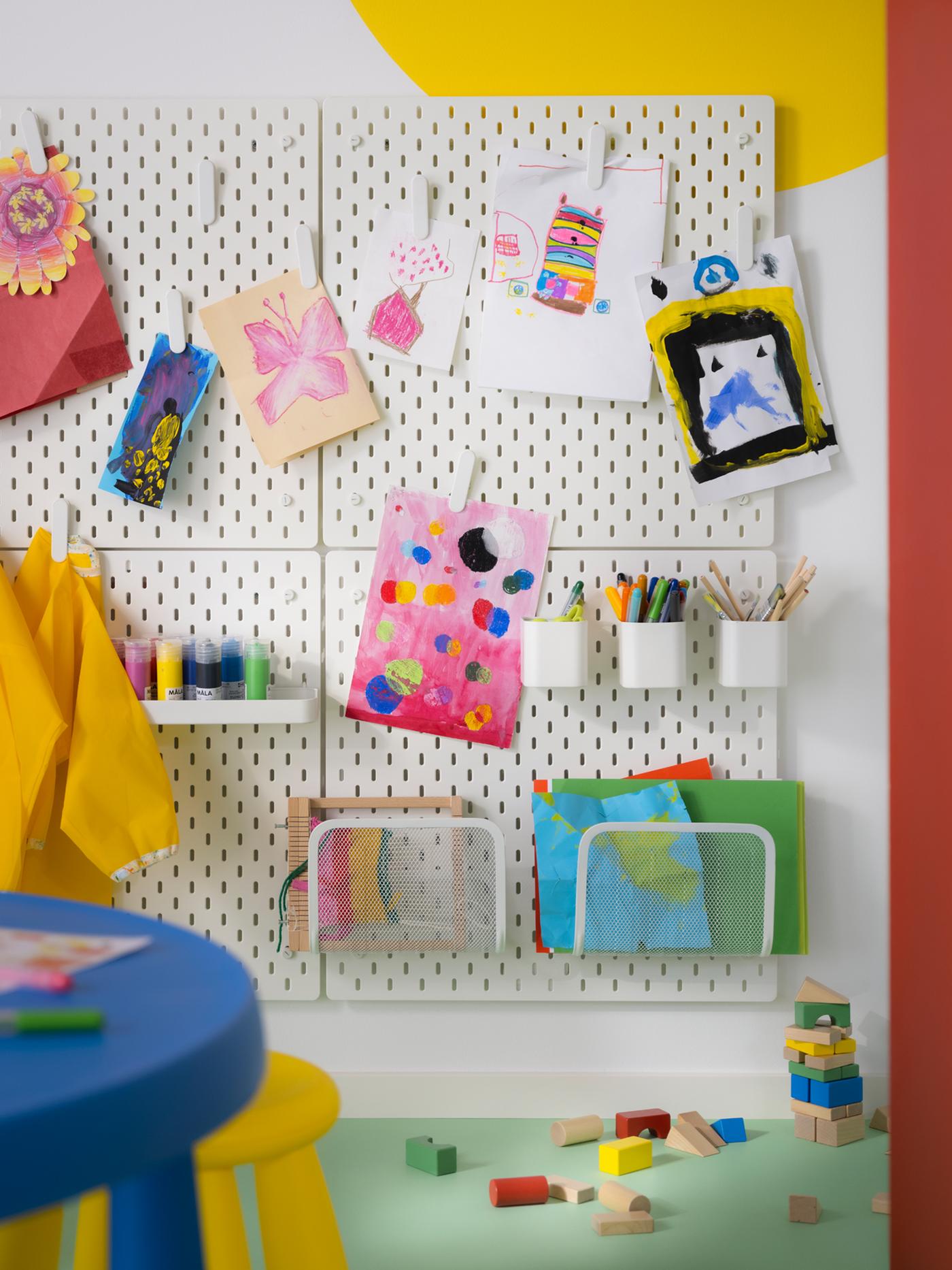 Dessins d’enfants exposés sur quatre panneaux perforés SKÅDIS. Les fournitures artistiques sont rangées sur une tablette et dans des récipients et range-revues de la série SKÅDIS.