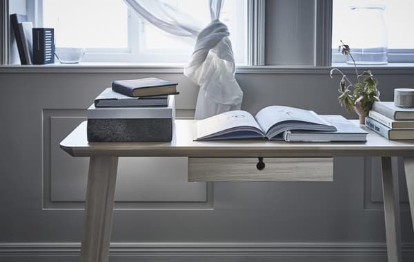 Detail of a desk with open books on top, in front of a window with a knotted curtain.