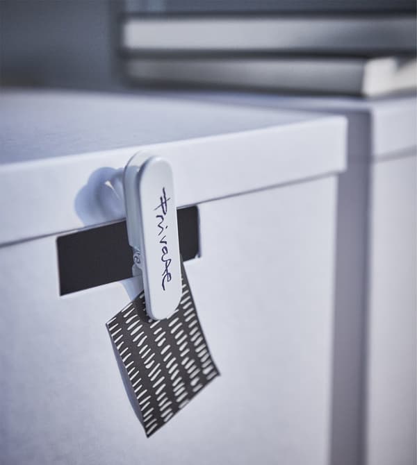 Detail of white IKEA TJENA storage boxes with pegs and labels telling us what's inside.
