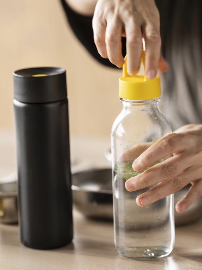 Detail rukou zavírajících průhlednou láhev na vodu s žlutým víčkem IKEA VATTENKRASSE, vedle stojí černá termoska IKEA EFTERTRÄDA na světlém stole.