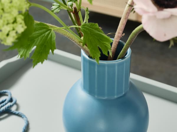 Detail vázy OMFÅNG v modré barvě na okdládacím stolku, ve váze jsou zelené rostliny