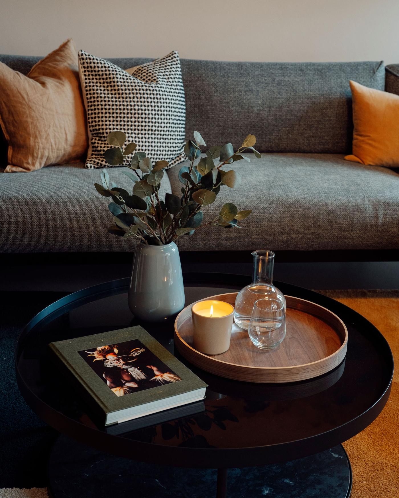 Detailansicht eines runden Couchtischs mit einem Holztablett einer Vase mit Zweigen und einem Buch darauf. Dahinter steht ein Sofa mit Zierkissen. 