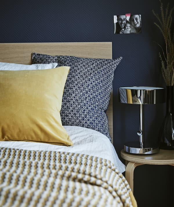 Detalle da esquina dunha cama, mesa de noite e lámpada xunto a unha parede azul escuro.