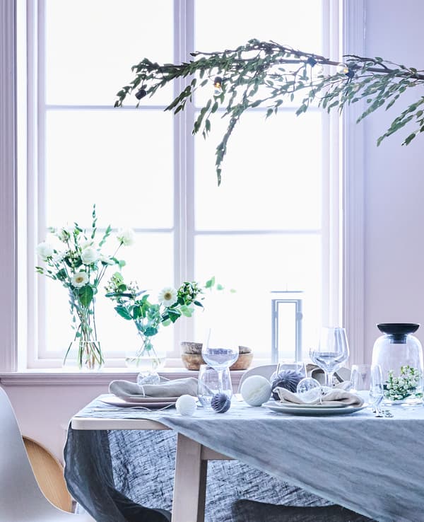 Detalle da mesa da sala con pezas de cristalería e decoracións en branco e gris xunto cun mantel de liño gris.