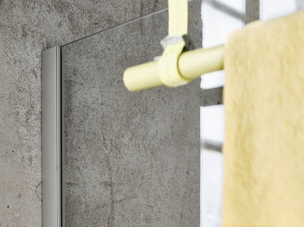 Detalle de la mampara de ducha OPPEJEN unida a una pared de hormigón gris con una barra amarilla y una toalla colgada en primer plano.