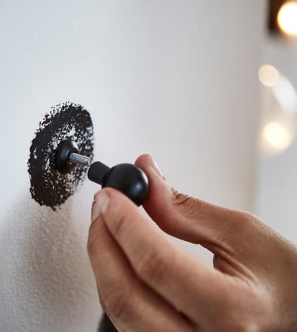 Detalle de una mano que sostiene un tirador de cocina BAGGANÄS negro, uniéndolo a uno de los puntos negros en la hoja de madera contrachapada blanca.