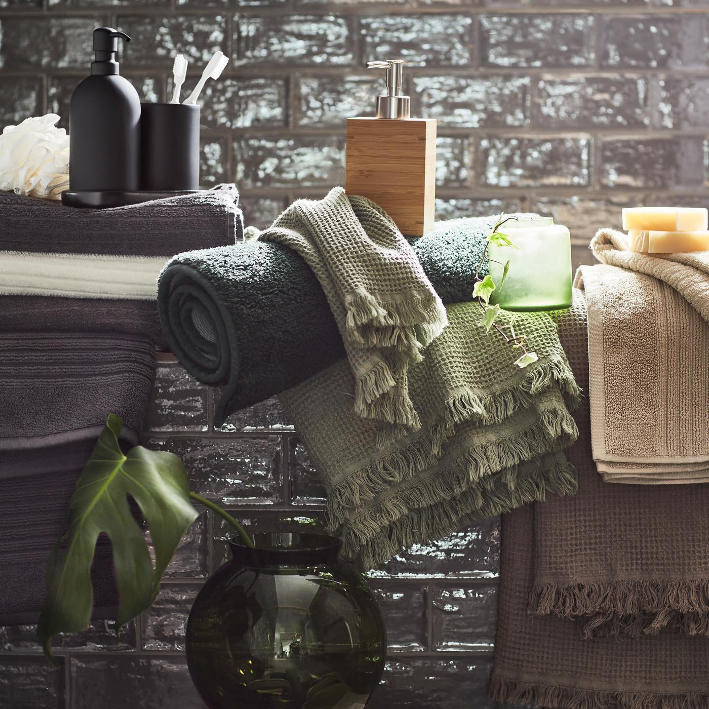 Detalle de varias toallas VALLASÅN en verde apagado, gris y beige en un baño con azulejos en gris oscuro.