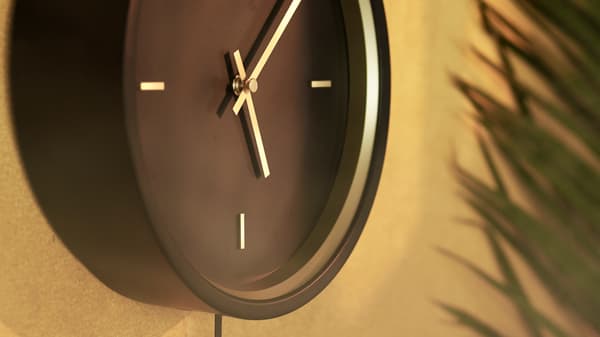 Detalle del reloj de pared STURSK negro con detalles y manecillas doradas, colgado en una pared de color marrón dorado.