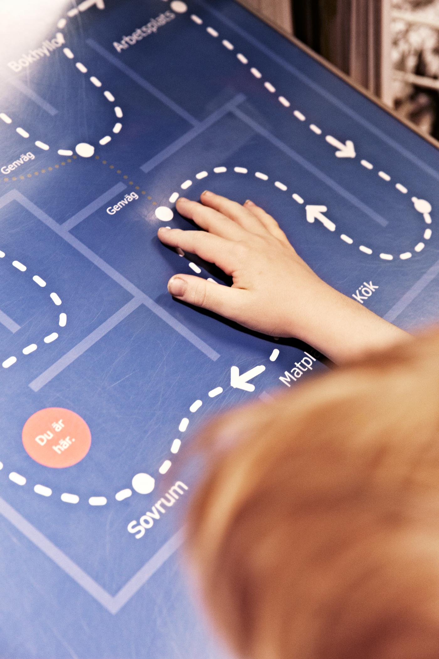 Dete dodiruje mapu sa belim isprekidanim linijama koje prikazuju putanju kroz robnu kuću IKEA, uz oznake na švedskom jeziku.