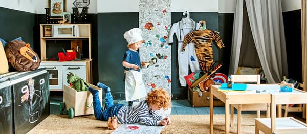 Deti sa hrajú v izbe s dvomi malými stolmi a stoličkami, tabuľou, miniatúrnou kuchyňou a vlajkami.