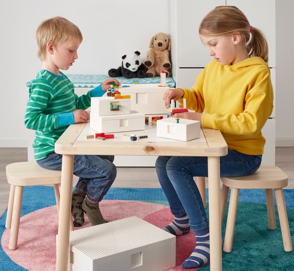 Deti sediace za dreveným stolom hrajúce sa s dielmi LEGO pripevňujúce minifigúrky LEGO k dvom bielym škatuliam BYGGLEK.