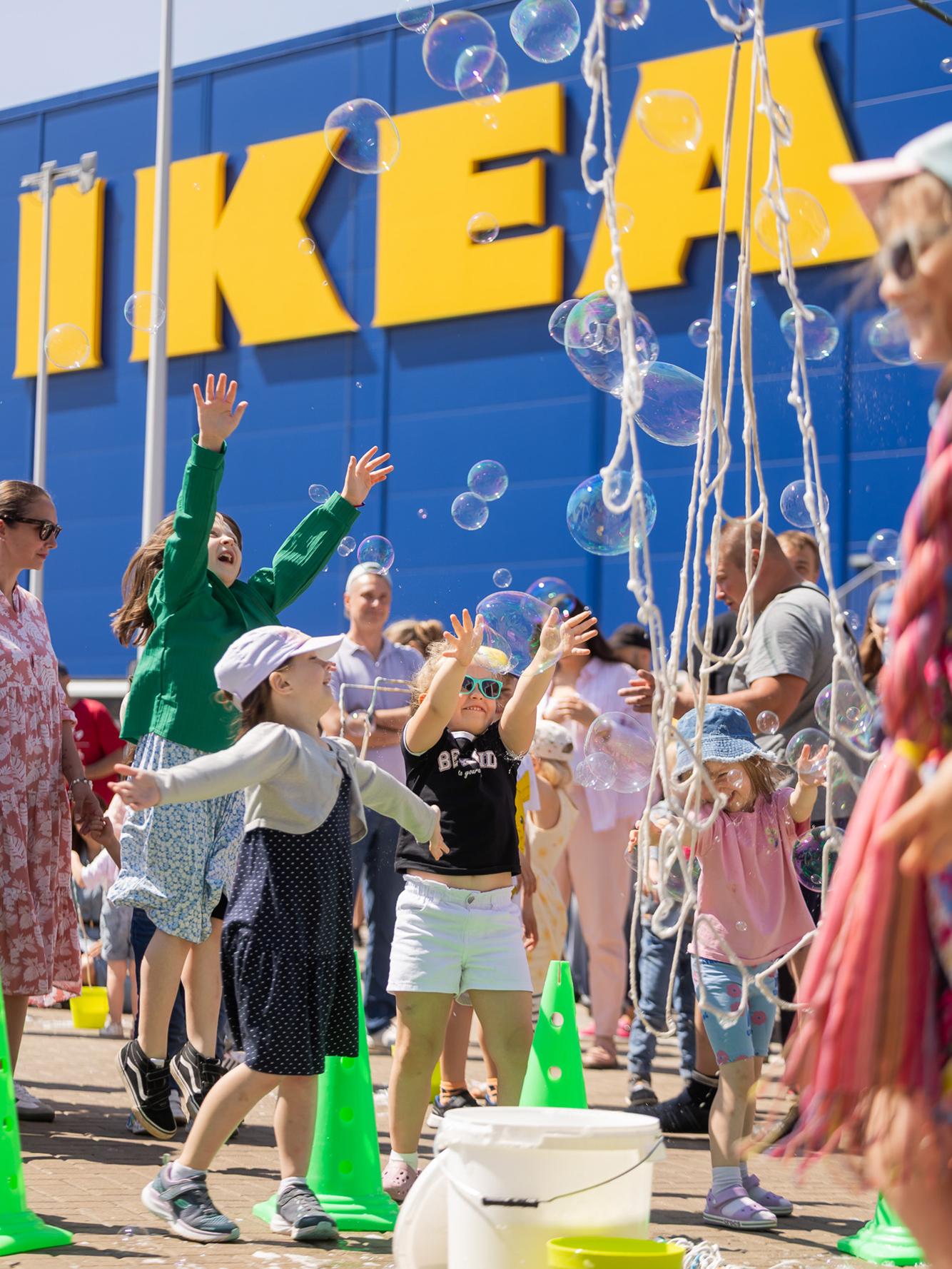 Дети запускают большие мыльные пузыри перед магазином IKEA. На заднем плане на синей стене жёлтыми буквами написано «IKEA».