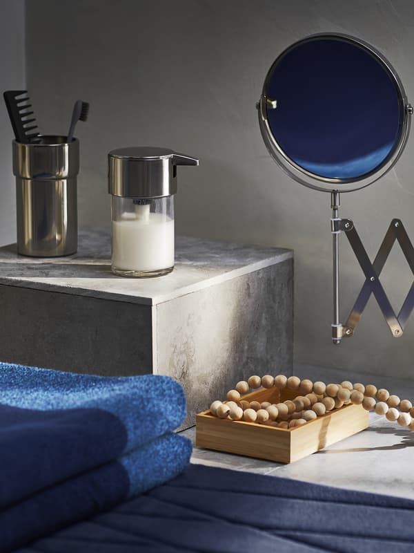 Dettaglio di un bagno con alcuni asciugamani piegati blu, una collana, un portaspazzolino e un dispenser. A destra, compare uno specchio FRÄCK.
