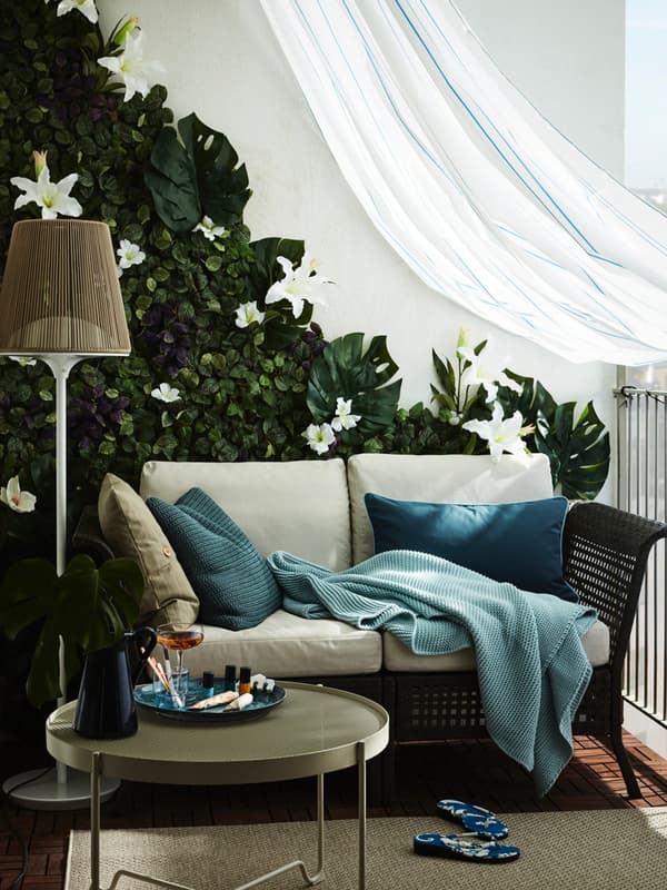 Dettaglio di un balcone con tenda leggera. Si vede un divano da esterno con grossi cuscini, di fronte un tavolino KROKHOLMEN e accanto una lampada da terra.