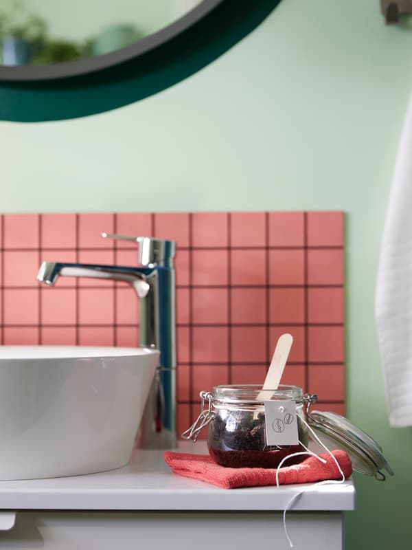 Dettaglio di un lavabo di colore bianco con rubinetto. Accanto, un vaso in vetro aperto con una palettina all'interno.