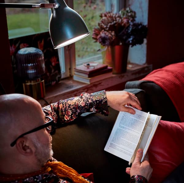Dettaglio di un uomo che legge un libro seduto su un divano. C'è una lampada che illumina il libro e accanto a lui una finestra con diversi oggetti.