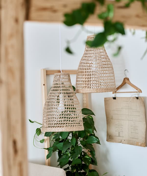 Deux abat-jour pour suspension tissés dans un matériau naturel sont suspendus à côté d’une plante verte feuillue et des feuilles de papier sont accrochées à une pince.