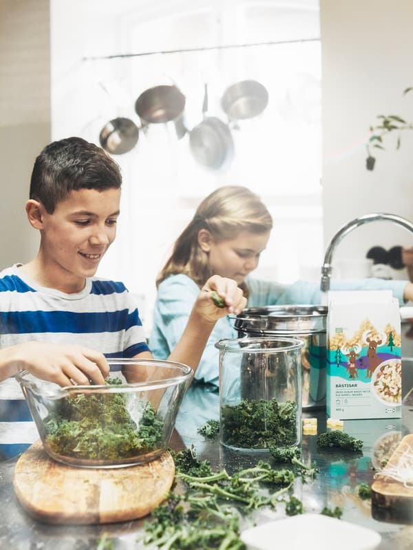 Deux adolescents préparent des pâtes et des légumes sur un plan de travail sur lequel sont posés un bol en verre, un bocal, une passoire, et un paquet de pâtes BÄSTISAR.