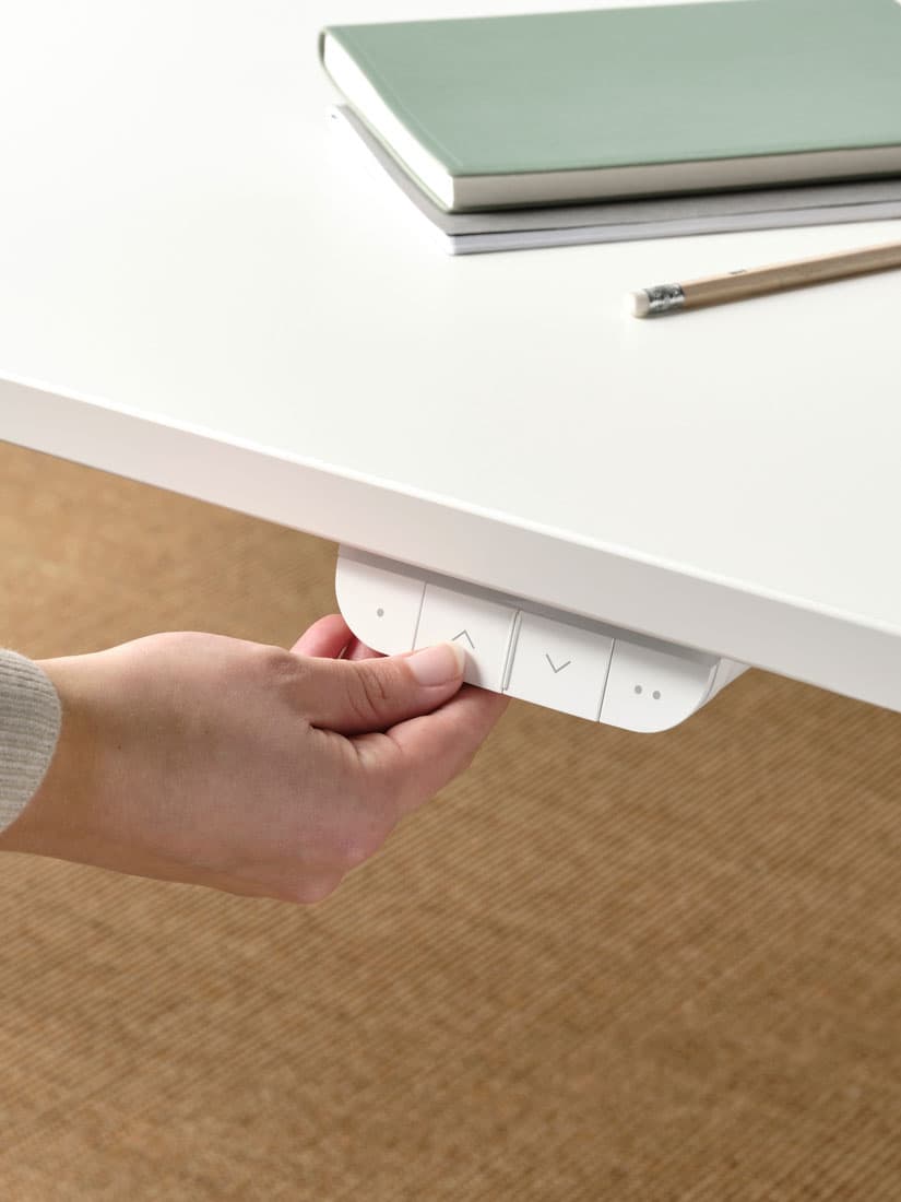 Deux bureaux MITTZON l’un à côté de l’autre, dont un haut avec tabouret assis-debout LIDKULLEN beige. Une femme est assise sur une chaise HATTEFJÄLL.