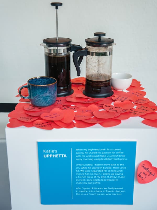 Deux cafetières à piston UPPHETTA sont en présentation avec une tasse bleue et une tasse blanche. Plusieurs notes autocollantes en forme de cœurs sont collées sur le présentoir.