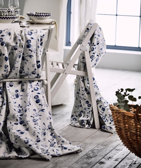 Deux chaises pliantes drapées d’un tissu blanc à motif floral bleu BLÅGRAN à proximité d’une table joliment décorée.