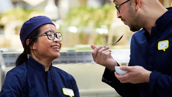 Deux collaborateurs IKEA habillés en uniformes bleu foncé qui ricanent en mangeant une collation.
