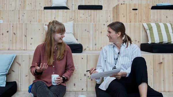Deux collaboratrices IKEA dans une tenue décontractée qui discutent en profitant d’un café, assises sur des marches en bois.