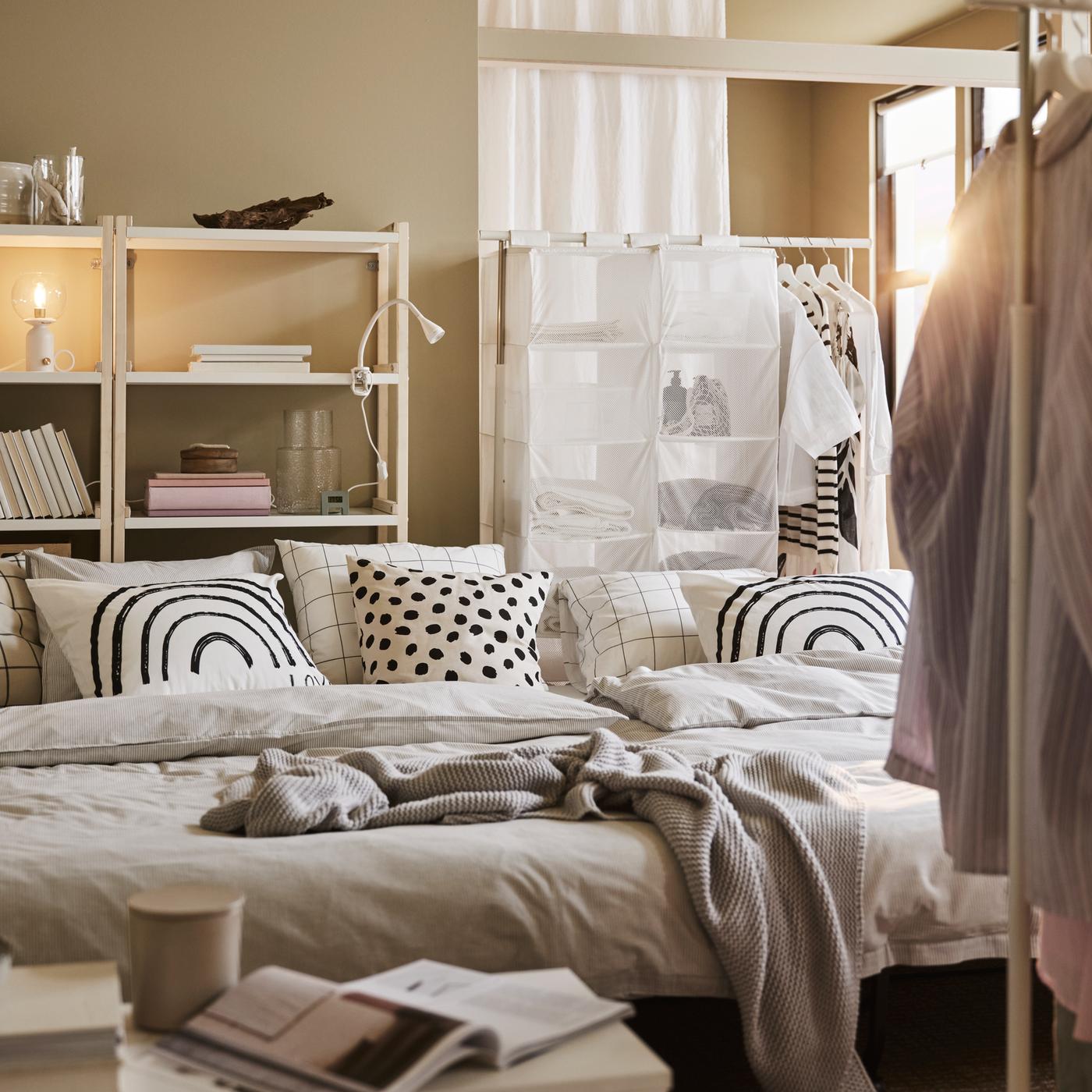 Deux convertibles LYCKSELE LÖVÅS avec du linge de lit gris et des oreillers à motifs, plus une table basse blanche et des portants pour vêtements blancs.