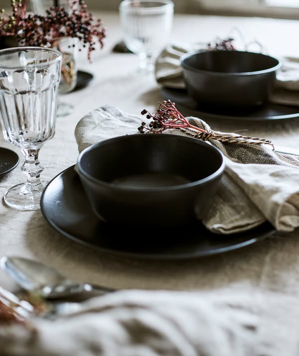 Deux couverts dressés sur une nappe en lin, chacun composé d’un bol gris-bleu sur une assiette avec un verre et une serviette nouée.