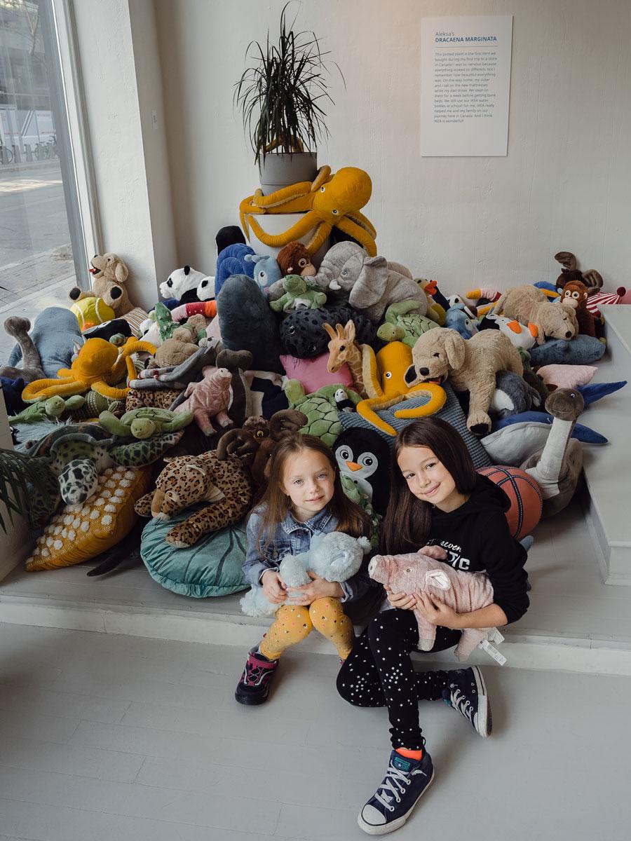 Deux enfants sont assis devant un présentoir exposant un grand nombre de peluches et une plante placée au centre du présentoir.