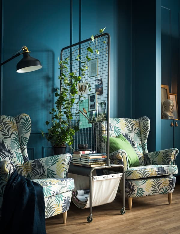 Deux fauteuils séparés par une cloison avec plante, magazine et rangement pour livres.