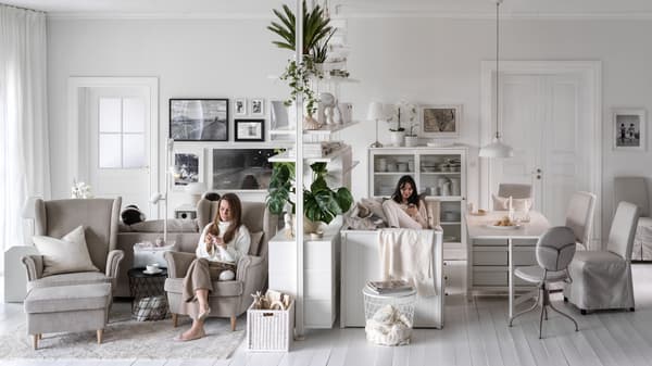 Deux femmes assises, une dans un fauteuil STRANDMON beige, de chaque côté d’un élément de rangement ELVARLI dans un grand séjour aéré.