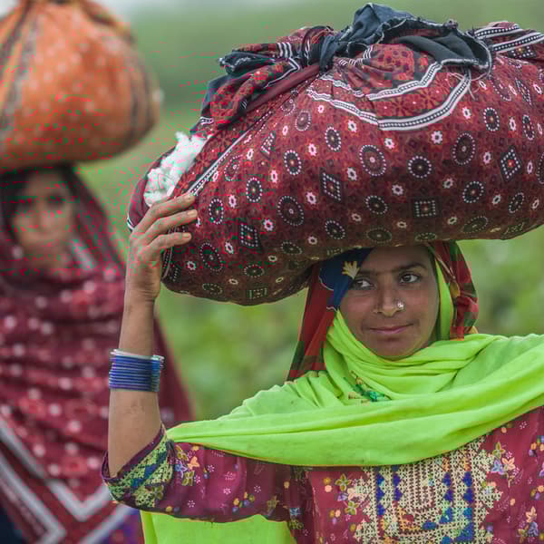 Deux femmes portant des vêtements colorés et transportant sur leur tête de gros sacs en coton, remplis de fibres de coton récoltées.