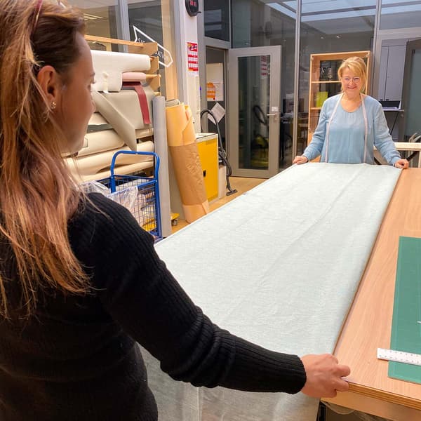 Deux femmes se tiennent face à face à une grande table de tailleur et lissent une grande feuille de tissu gris dans laquelle un rideau doit être cousu.