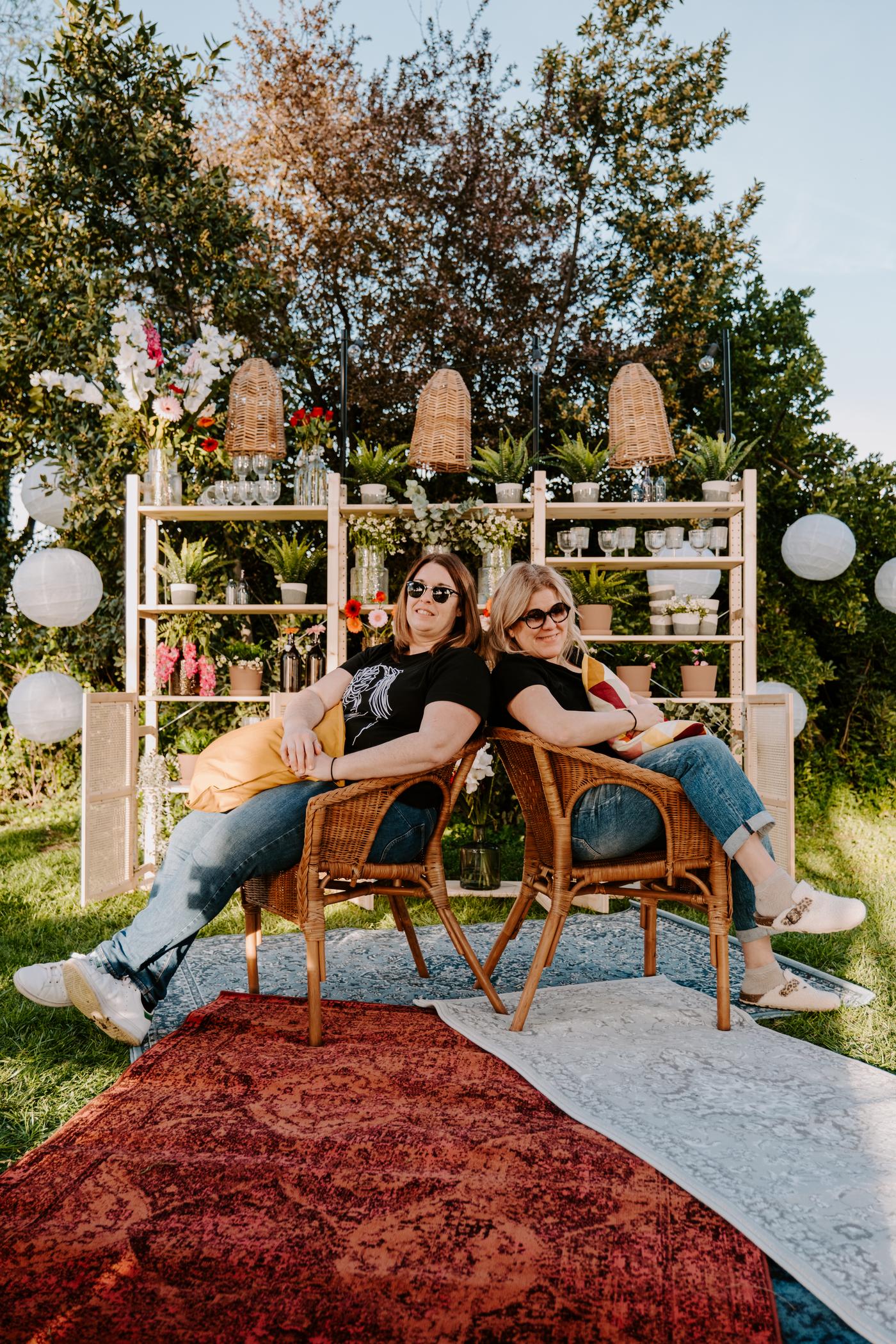 Deux femmes sont assises dos à dos sur des chaises dehors devant des étagères remplies de décorations estivales
