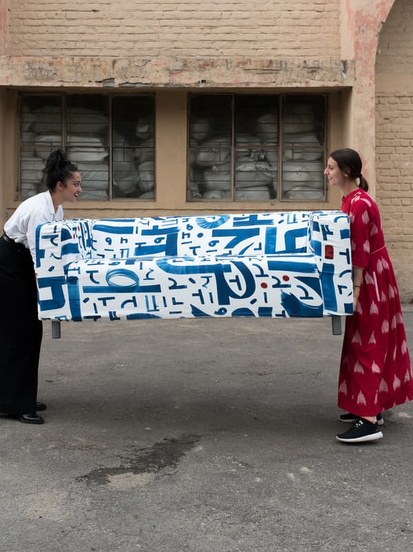 Deux femmes tenant les deux extrémités d’un canapé avec une housse en tissu blanc et bleu sur un sol asphalté, devant un entrepôt en briques.